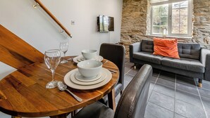 Un coin repas avec une table en bois dressée pour quatre personnes, un canapé gris agrémenté d’un coussin orange et un mur en pierre avec une fenêtre.