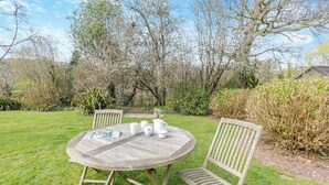 Une table ronde en bois, entourée de chaises, se trouve au milieu d’un jardin verdoyant agrémenté d’arbres et d’arbustes.