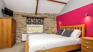 Une chambre avec un mur en pierre, un lit en bois, une fenêtre décorée avec le mot « AMOUR », un téléviseur fixé au mur et un mur d’accent de couleur rouge.