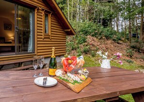 Ein Holztisch ist gedeckt mit Champagner, Meeresfrüchten und Blumen, im Hintergrund ist eine Hütte zu sehen.