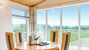 A dining area with a round table, four chairs, a vase with flowers, and a view of a green landscape through large windows.