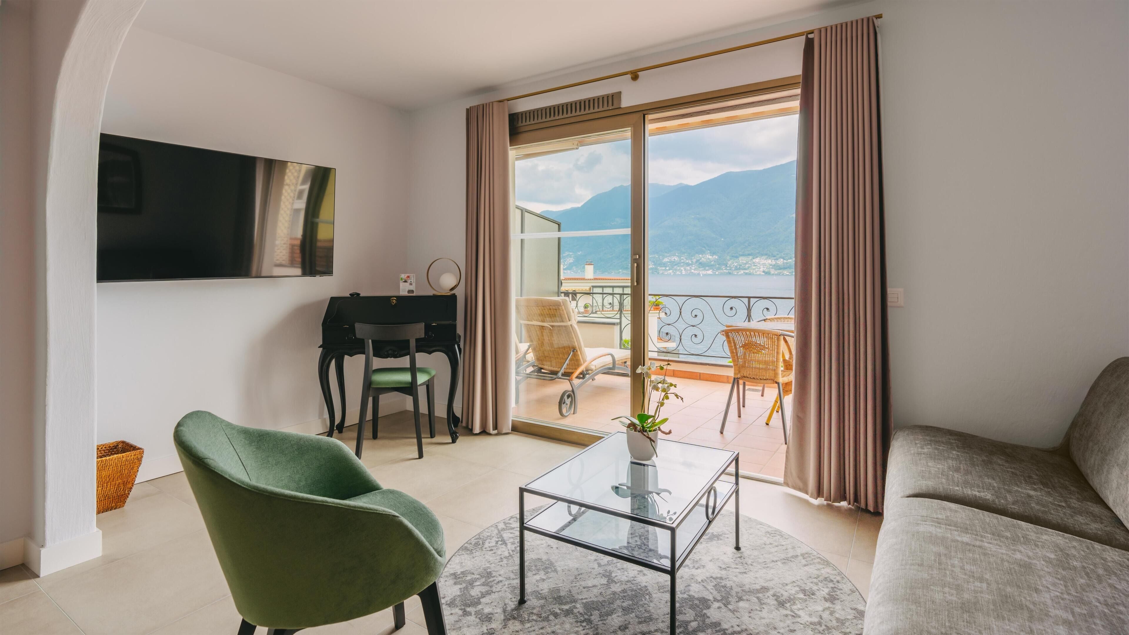Una sala de estar moderna con un sillón verde, un sofá, una mesa de centro de vidrio y un escritorio con una silla. Un ventanal ofrece vistas al mar y a las montañas.