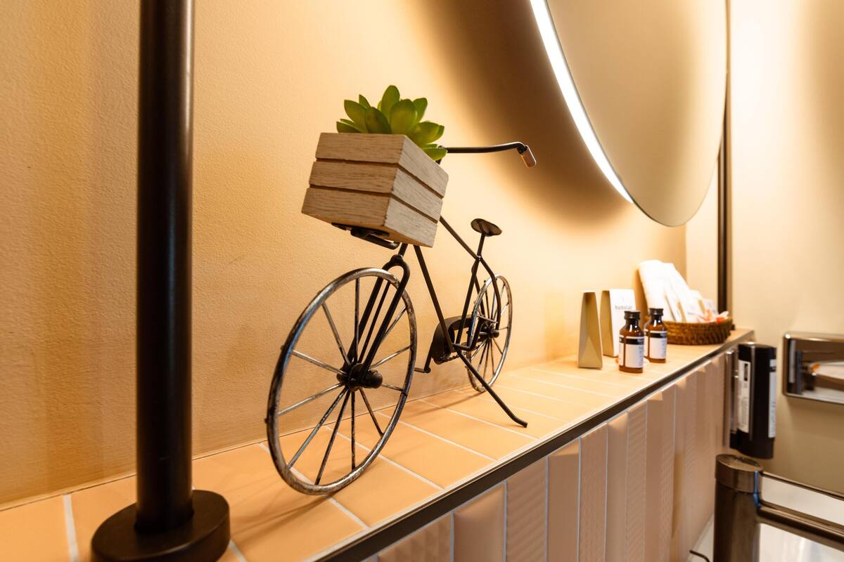 Una bicicleta con una cesta que contiene libros y una planta, colocada sobre una encimera de baldosas con un espejo y artículos de aseo.