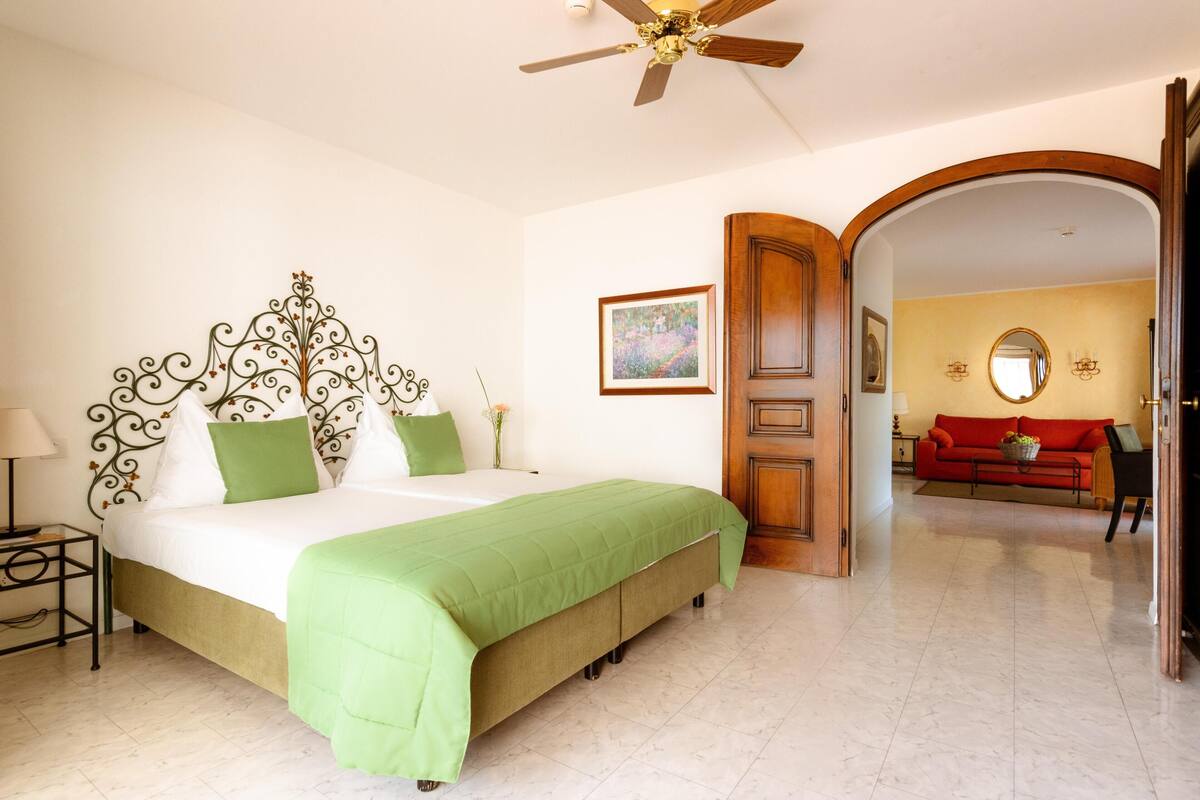 Un dormitorio con cama, mesita de noche y ventilador de techo, y un sofá rojo en la sala contigua.