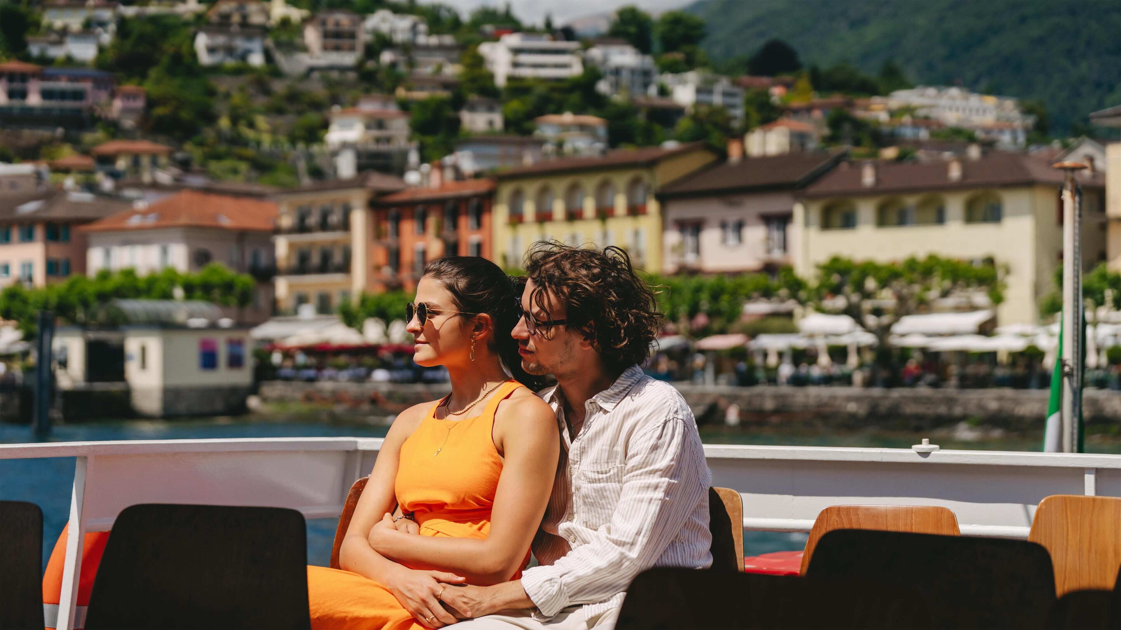 Un couple est assis sur un bateau, avec une vue sur une ville côtière et des collines en arrière-plan.