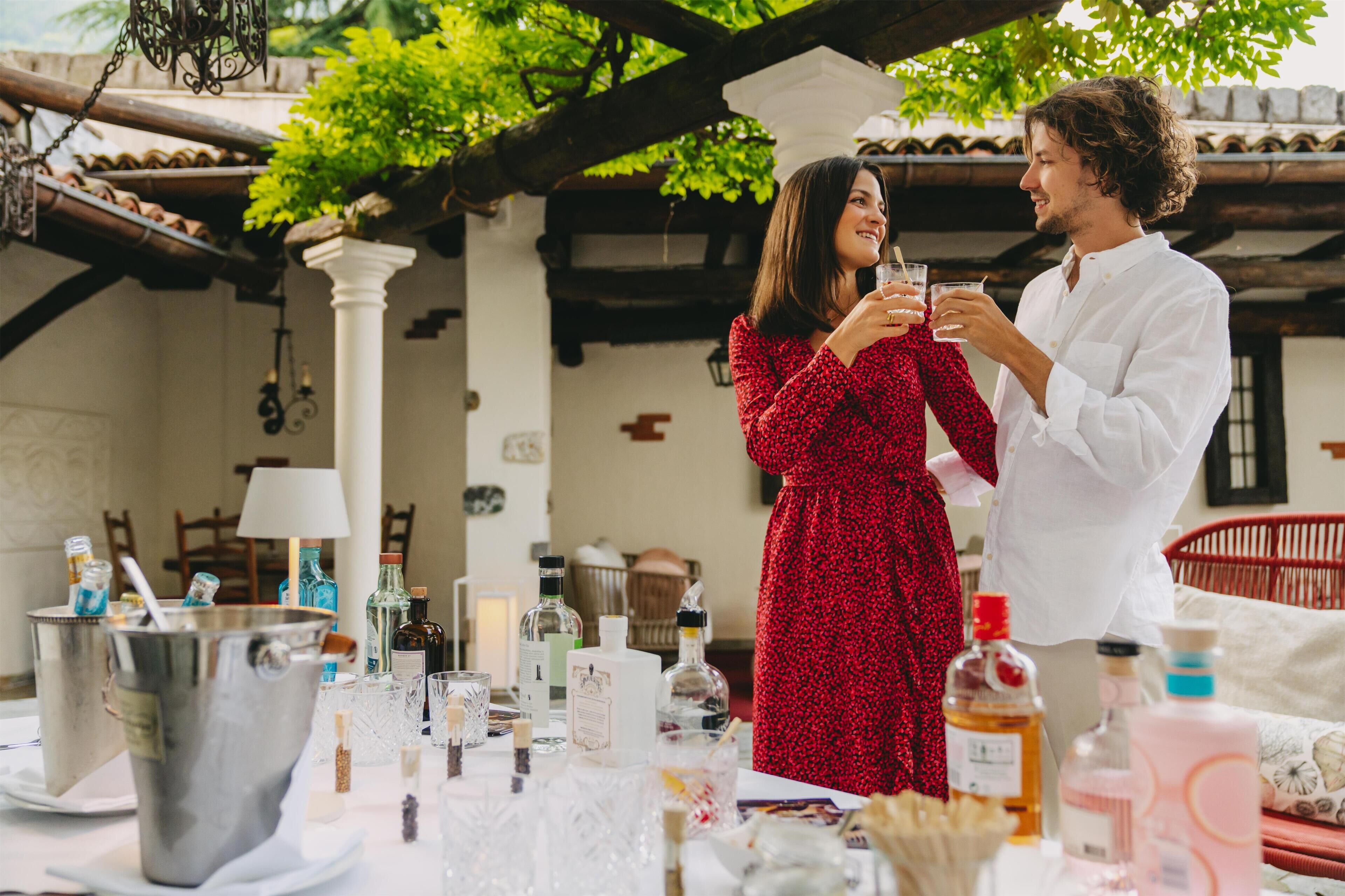 A couple toasting with drinks at an outdoor table with a tiled roof, surrounded by bottles and glasses.