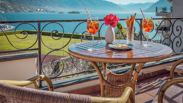 Una mesa redonda con un vaso de bebida, un jarrón con flores y un plato de comida en un balcón con vistas a un lago y montañas al fondo.