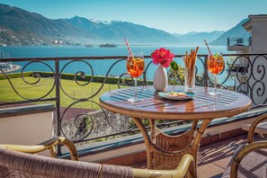 Una mesa redonda con un vaso de bebida, un jarrón con flores y un plato de comida en un balcón con vistas a un lago y montañas al fondo.