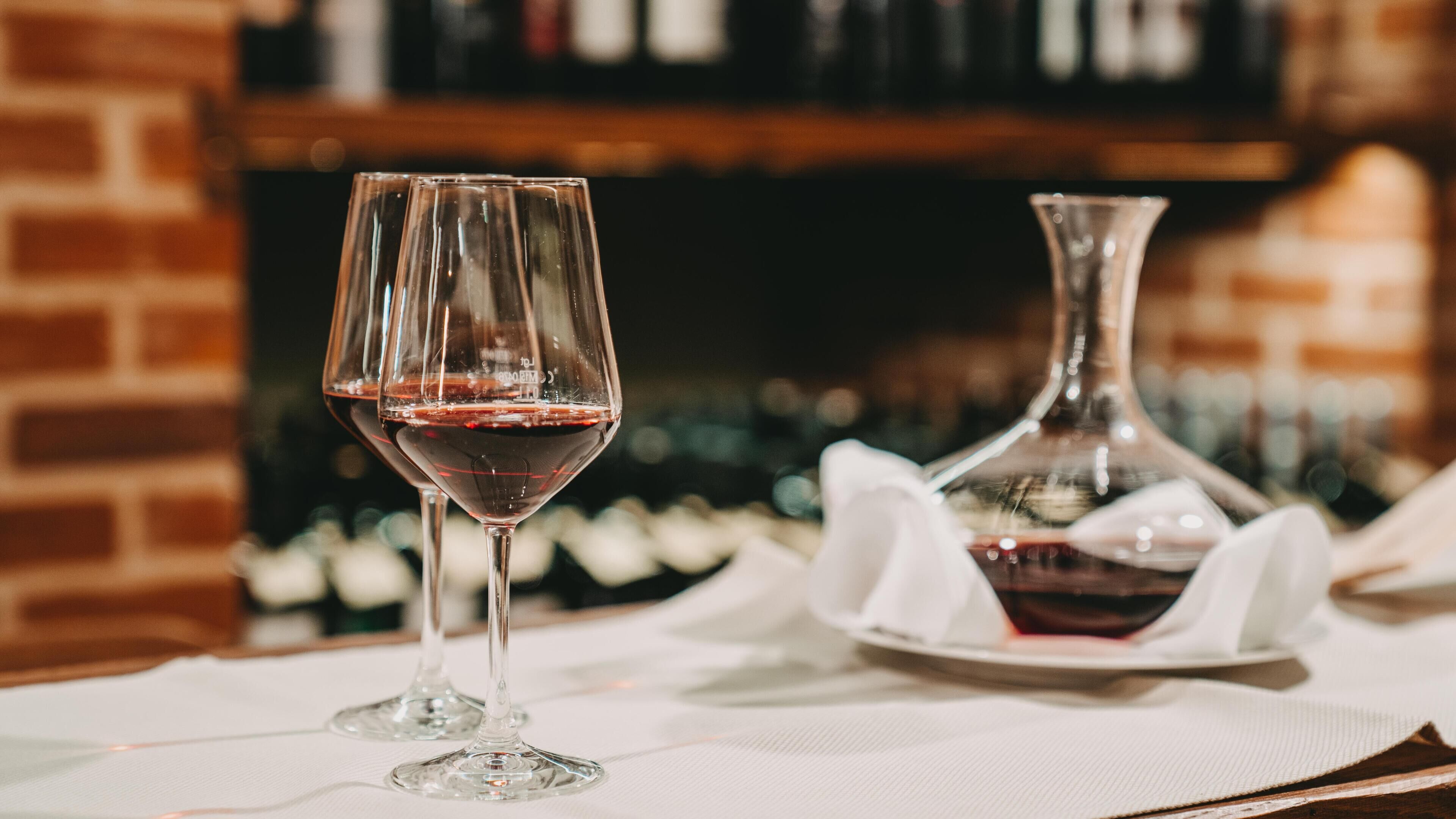 Two wine glasses on a table with a bottle of wine and a decanter in the background.