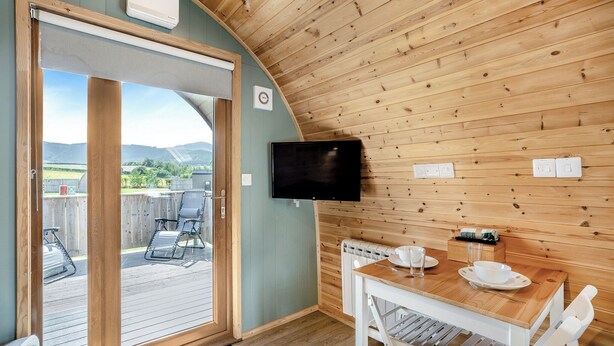 A wooden cabin with a sliding glass door leading to an outdoor deck, a television mounted on the wall, and a dining table with chairs. - 0 bedroom accommodation in Great Busby (Great Busby)