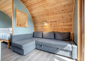 A cozy room with a wooden ceiling, a grey sofa, a bed with a striped duvet, and a wall-mounted light fixture.