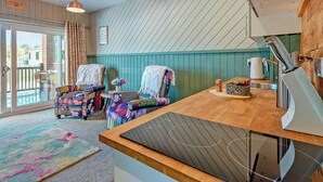 Een gezellige kamer met een houten tafel, stoelen met kleurrijke bekleding, een koffiezetapparaat en uitzicht door een glazen deur op een buitenruimte.