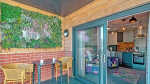Een gezellig interieur met een bakstenen muur, een groene wand met planten, een glazen deur die leidt naar een keuken, en een klein zithoekje met stoelen en tafels.