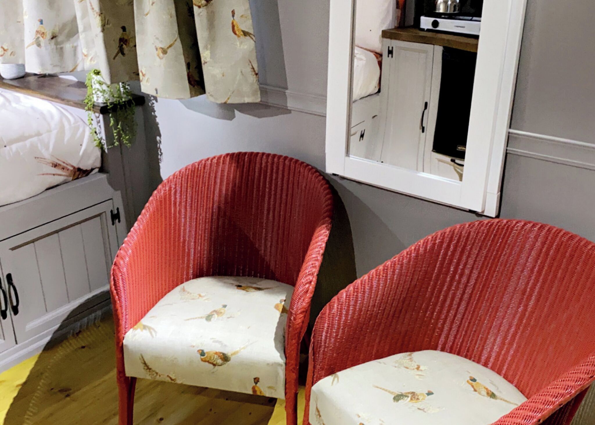 A room with two red wicker chairs with patterned cushions, a white cabinet with a mirror, and a window with curtains.