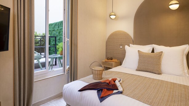 A hotel room with a bed, a television, a small table with a chair, a wicker basket, and a view of a balcony with greenery. - 1 bedroom apartment with private terrace - 44m2 (Paris)