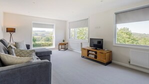 Una sala de estar moderna con un sofá gris, un televisor sobre un mueble de madera y ventanas amplias con vistas a zonas verdes.