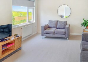 Ett vardagsrum med en grå soffa, en tv-apparat på ett träställ, ett stort fönster med utsikt över åkrar och en rund spegel på väggen.