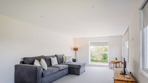 Una sala de estar moderna con un sofá seccional gris, una mesita de madera, una lámpara de pie y un amplio ventanal con vista a la vegetación.