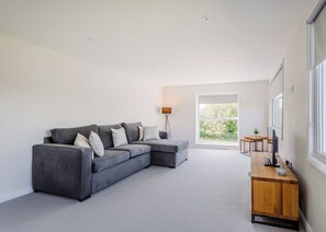A modern living room with a grey sectional sofa, a wooden side table, a floor lamp, and a large window with a view of greenery. - 1 bedroom accommodation in South Molton, nr Barnstaple (South Molton, nr Barnstaple)