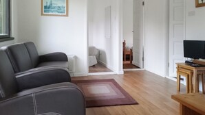 A living room with a sofa, a TV on a wooden stand, a framed picture on the wall, and a door leading to another room. - 2 bedroom accommodation in North Tamerton, Bude (North Tamerton, Bude)