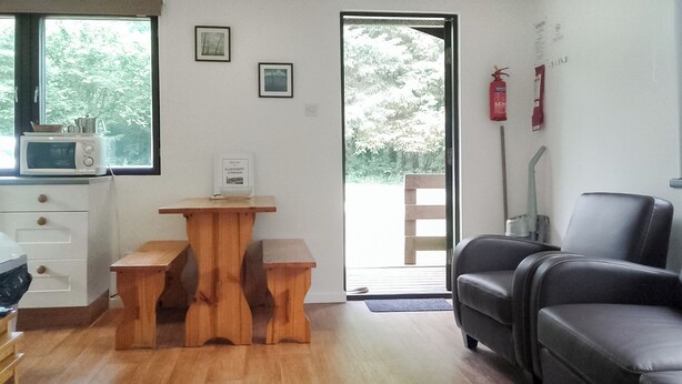 A living room with a wooden table, a black leather sofa, a microwave, and a door leading outside. - 2 bedroom accommodation in North Tamerton, Bude (North Tamerton, Bude)