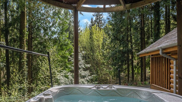 Een hottub in een bebost gebied, met een houten terras en een prieel.