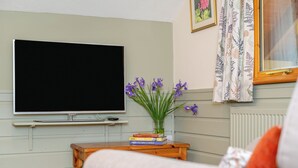 Een woonkamer met een flatscreen televisie, een klein houten bijzettafeltje met boeken, een vaas met bloemen en een bank.