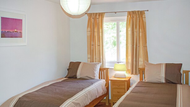 A bedroom with two single beds, a bedside table with a lamp, and a window with curtains. - Ground floor apartment with one double, one twin and one with bunks. (Dawlish Warren)