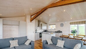 A cozy cabin interior with two grey sofas, wooden flooring, a kitchen with white cabinets, and a dining area with a table and chairs. - Field View Lodge (Pet) (Bretby, Derbyshire)
