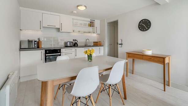 Una cocina moderna con armarios blancos, una mesa de comedor con sillas, una mesita auxiliar de madera y un reloj de pared.