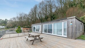Una terraza de madera con una mesa de picnic, un edificio con paredes de vidrio y vistas a árboles y un edificio lejano.