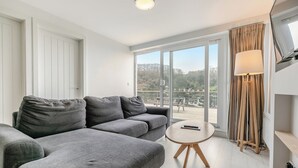 Uma sala de estar moderna com um sofá cinzento, uma mesa redonda pequena, um abajur e uma grande porta de vidro deslizante que dá para um deck externo.