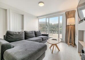 A modern living room with a grey sofa, a small round table, a floor lamp, and a large sliding glass door leading to an outdoor deck. - Sea Breeze Premier apartments are perfect for a getaway with friends. (Millendreath, Looe)