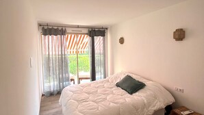 A bedroom with a large bed, a small bedside table, a window with curtains, and a view of a balcony.