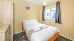 Un dormitorio con cama, armario, ventana con cortinas, un cuadro pequeño en la pared y una lámpara en el techo.