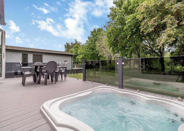 Un jacuzzi en una terraza de madera con mobiliario exterior y vista a una zona arbolada.