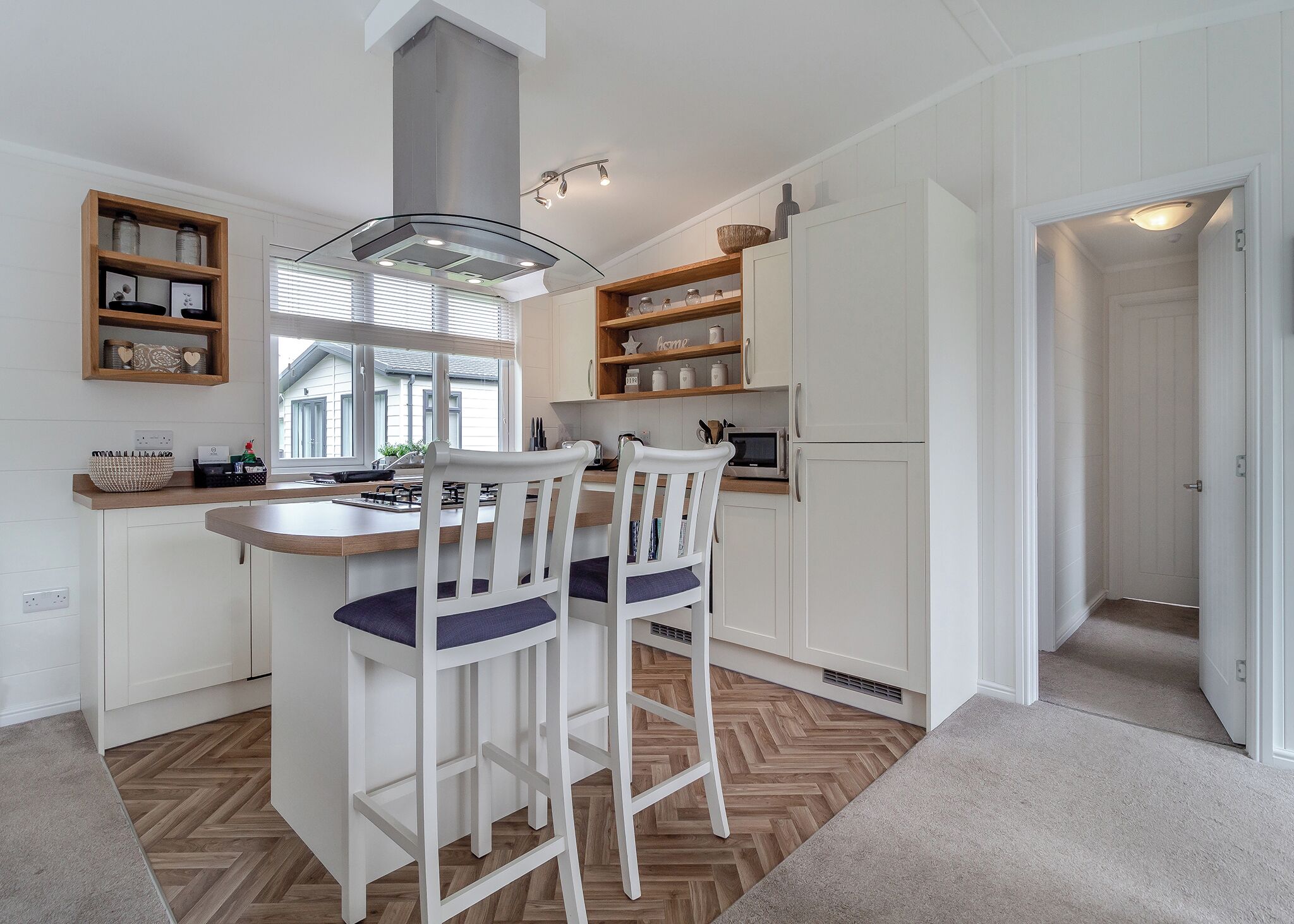 A modern kitchen with white cabinetry, a central island with bar stools, a stainless steel range hood, and a window providing natural light.