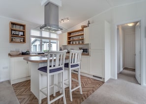 Una cocina moderna con armarios blancos, una isla central con taburetes de bar, una campana extractora de acero inoxidable y un ventanal que proporciona luz natural.