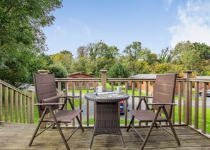 A deck with wicker chairs and a glass table, overlooking a green landscape with trees and a building.