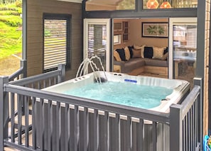 A hot tub with a wooden deck and a view of a living room with a sofa and cushions. - Riverside Family Pod HT (Snowdonia)