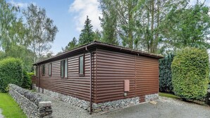 Ein Blockhaus mit Steinfundament, einem Schotterweg und einem gepflegten Garten mit Hecke und Bäumen.