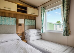 A compact RV bedroom with two beds, a wooden headboard, a window with curtains, and a small shelf unit.