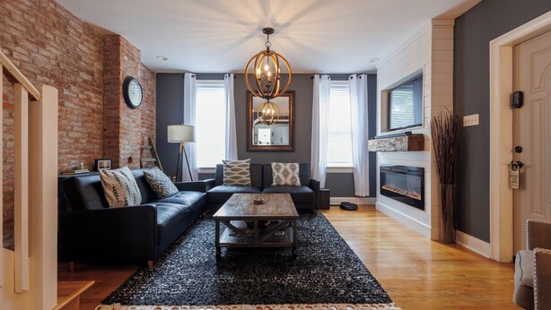 Een woonkamer met een zwarte leren bank, een houten salontafel, een open haard en een met baksteen beklede muur.