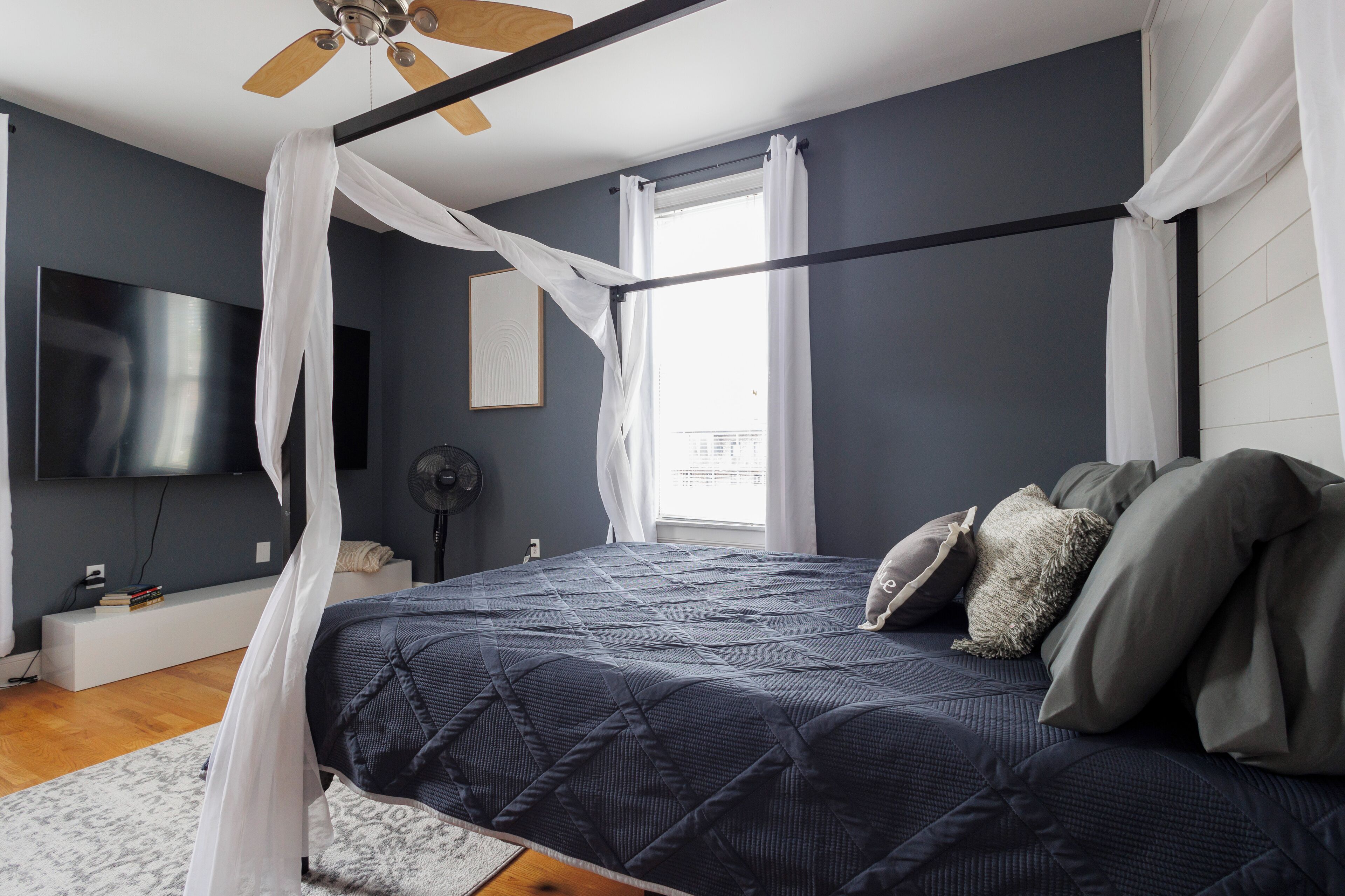 Un dormitorio con cama, ventilador de techo, televisión y ventana con cortinas.