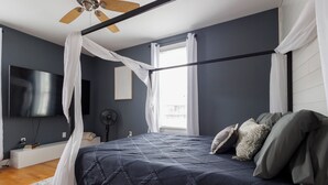 Een slaapkamer met een bed, een plafondventilator, een televisie en een raam met gordijnen.