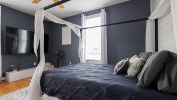 Un dormitorio con cama, ventilador de techo, televisión y ventana con cortinas.