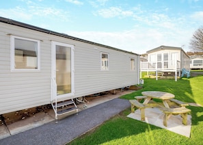 A mobile home with a front porch, a small patio with a table and chairs, and a grassy yard.