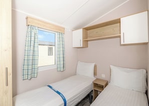 A compact room with two single beds, a window with plaid curtains, a small wooden shelf, and a white wardrobe.
