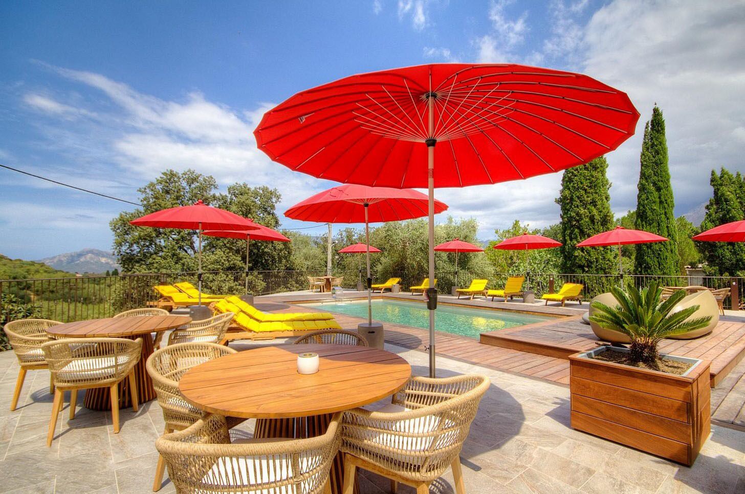A poolside area with wicker furniture, red umbrellas, and a view of mountains.