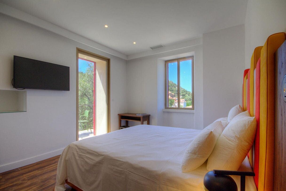 Un dormitorio con cama, televisor montado en la pared y dos ventanas con vistas a zonas verdes y edificios.